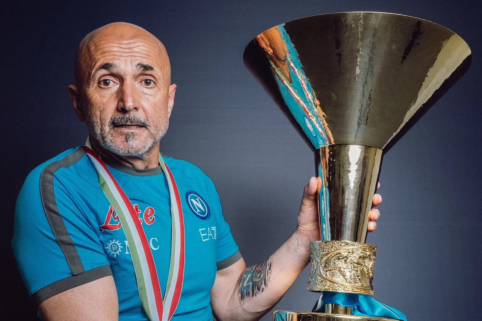 A man wearing a light blue Napoli jersey and an Italian flag-colored medal, holding a large golden trophy