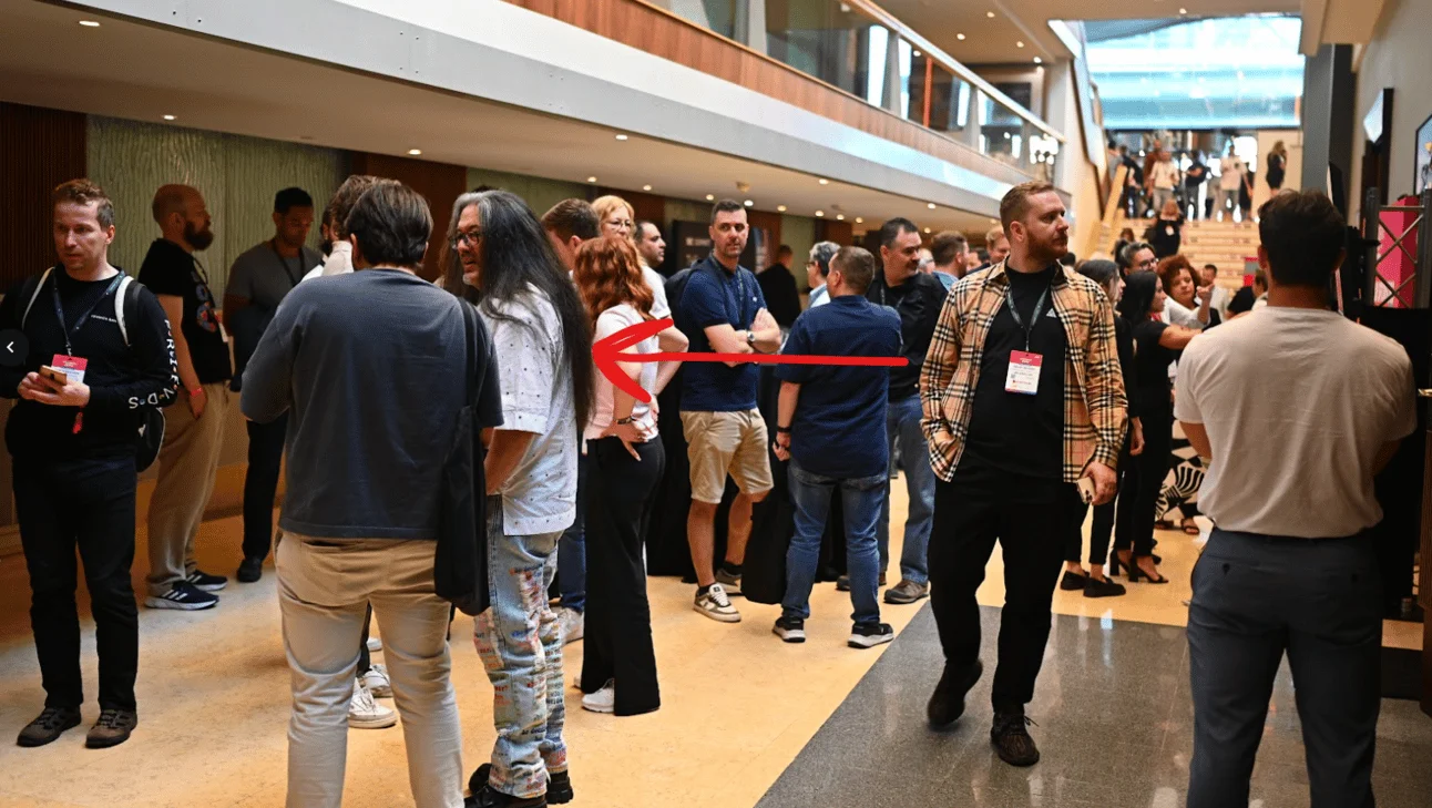 A crowded conference hallway with many people standing and talking in groups, with a red arrow drawn across the image